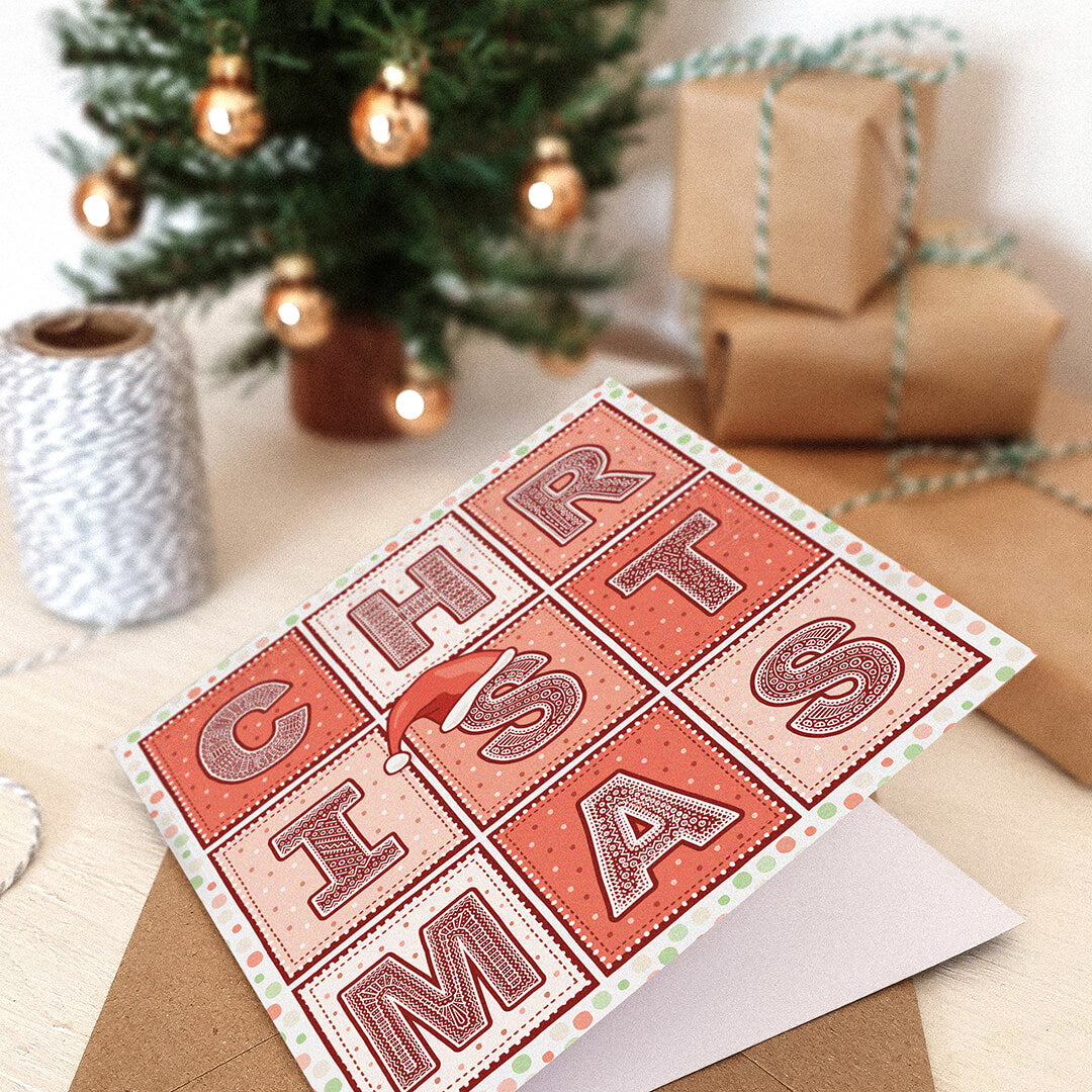 Christmas card with decorative text in a festive setting with a small tree and wrapped gifts.